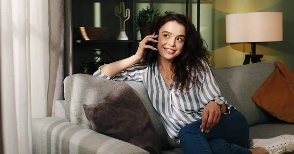 Расслабленный довольный кавказский молодой радостный симпатичная женщина разговаривает по телефону, сидя на диване в одиночестве в доме с улыбкой на лице в мечтательном настроении, женщина говорит по мобильному телефону — стоковое фото