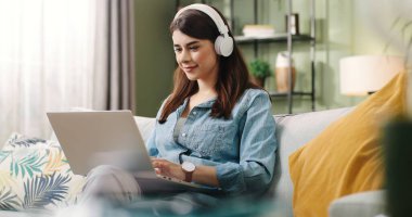 Genç ve güzel bir kız, evinde oturmuş dizüstü bilgisayarında yazı yazıyor ve beyaz kulaklıklarla müziğin keyfini çıkarıyor..