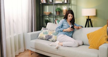 Güzel, genç, beyaz, neşeli bir kadın. Oturma odasında rahat bir koltukta oturuyor ve akıllı telefonuyla mesajlaşıyor. İnternetten sosyal ağ videoları izliyor.