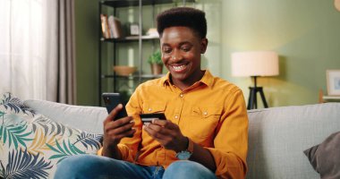Afro-Amerikalı neşeli genç yakışıklı erkek kanepede dinleniyor, internetten cep telefonuyla internetten kredi kartıyla alışveriş yapıyor. E-ticaret, portre konsepti