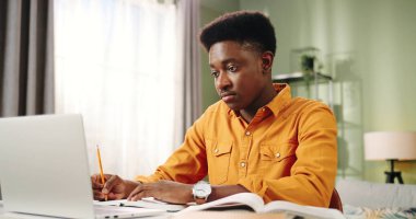 Portrait of serious concentrated young african american male student sitting at table in room at home and reading book writing in notebook learning on laptop. Distance studying concept. Self-education clipart