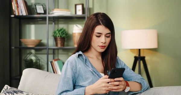 Красивая радостная улыбающаяся молодая кавказка, отдыхающая на удобном диване в гостиной и печатающая на смартфоне поиск в интернете смс с улыбкой на лице. Концепция коммуникации — стоковое фото