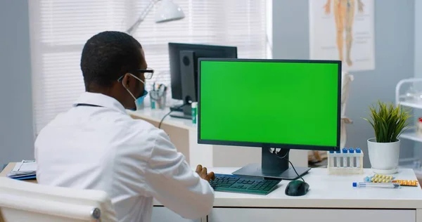 Back view of African American male physician sitting in hospital office and looking at green screen while working typing on computer. Medical concept, doctor browsing on monitor with chroma key