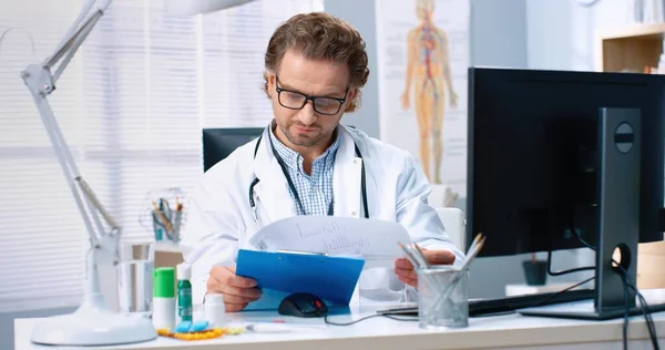 Portrait of Caucasian handsome adult experienced male physician specialist sitting at desk in cabinet checking medical documents and typing on computer in hospital, healthcare, work concept — Stock Photo, Image