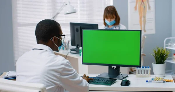 Back view of African American male physician sitting in hospital office and looking at green screen while working typing on computer. Medical concept, doctor browsing on monitor with chroma key