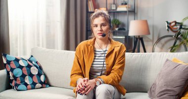 Evdeki oturma odasında kanepede otururken kameraya bakan güzel, genç, mutlu, beyaz kulaklı bir kadın. İnternet konferansında konuşan serbest çalışan kadın, e-öğrenim konsepti