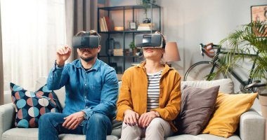 Kafkasyalı genç çift kanepede VR gözlük takarak oturuyor. Yenilikçi fütüristik teknolojiler kullanıyorlar. Evlerinde oturup el kol hareketleri yapıyorlar.