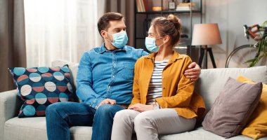 Tıbbi maskeli, mutlu, genç, beyaz bir çiftin portresi. Karantinada, kanepede oturmuş kameraya bakıp gülümsüyor. Aile zamanı, Coronavirus konsepti.