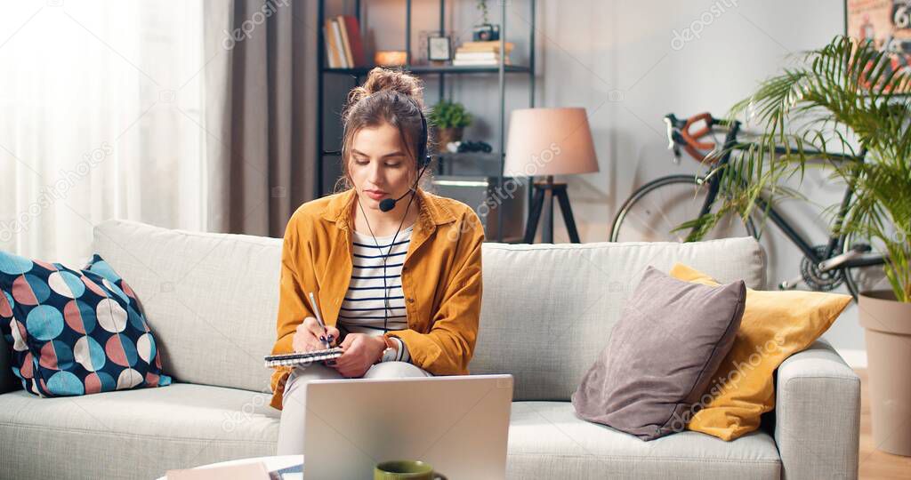 Beautiful young Caucasian woman in headset speaking on video call online on laptop computer while studying and writing down in notebook. Female freelancer working talking on online web conference