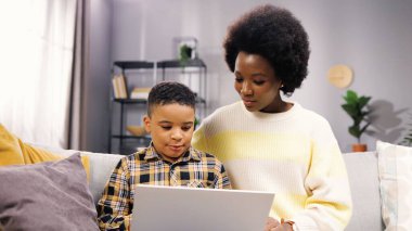 Mutlu Afro-Amerikan aile portresi güzel anne ve küçük oğul kanepede oturmuş evdeki dizüstü bilgisayarda geziniyorlar. Anne ve küçük çocuk bilgisayar eğitimi, e-öğrenme, eğitim konsepti