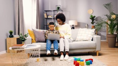 Afro-Amerikalı neşeli, genç, güzel bir anne ve kanepeye oturmuş dizüstü bilgisayarda yazı yazan sevimli bir çocuk. Küçük çocuk bilgisayarda çalışıyor annesiyle vakit geçiriyor, e-öğrenim konsepti