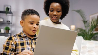 Afro-Amerikan neşeli, küçük sevimli oğlunun güzel annesiyle kanepede oturup dizüstü bilgisayarına göz atması. Annesiyle birlikte bilgisayar sörfü okuyan küçük çocuk, çocukluk konsepti.