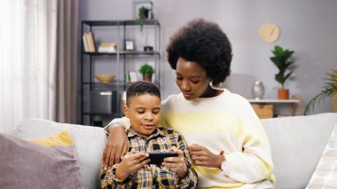 Genç anne ve babasıyla kanepede otururken akıllı telefondan yazı yazan tatlı Afro-Amerikan çocuk portresi. Teknoloji aleti, cep telefonu uygulaması, çocuk konsepti