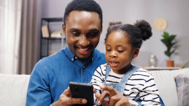 Neşeli, sevimli, Afro-Amerikan, küçük kız çocuğunun babasıyla oturma odasında kanepede oturması ve akıllı telefonları dinlemesi. Küçük kızına evde cep telefonu kullanmayı öğreten mutlu bir baba.
