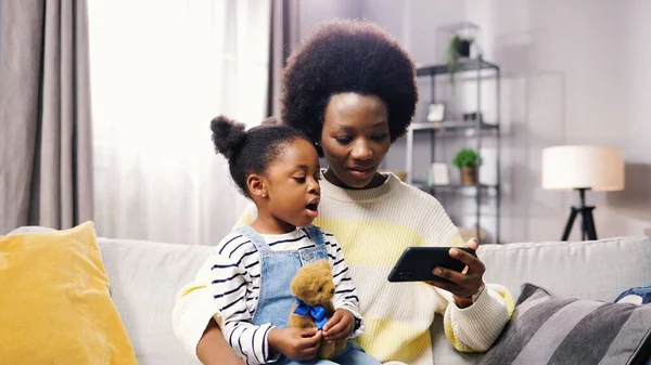 Portrait of happy African American family pretty mother and small adorable daughter sitting on sofa browsing online on smartphone watching cartoons. Mom and child searching internet on cellphone