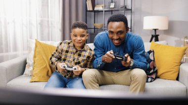 Afrikalı Amerikalı neşeli genç ebeveynin portresi. Küçük oğluyla eğleniyor. Kanepede oturuyor ve keyfi yerindeyken video oyunları oynuyor. Aile zamanı, oyun zamanı, oyun konsepti