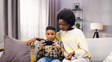 Genç anne ve babasıyla kanepede otururken akıllı telefondan yazı yazan tatlı Afro-Amerikan çocuk portresi. Teknoloji aleti, cep telefonu uygulaması, çocuk konsepti