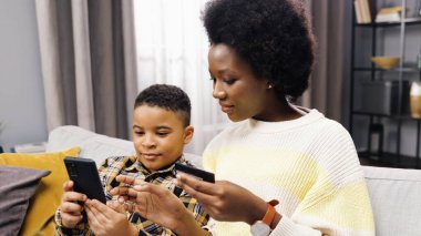 Genç ve güzel Afro-Amerikan annenin kanepeye oturup oğluyla banka kartı ve akıllı telefonu internetten alışveriş etmesi..