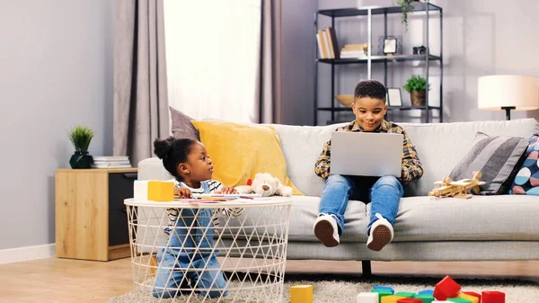 İki Afrikalı Amerikalı çocuk hafta sonlarını evde geçiriyor. Sevimli çocuk kanepede oturup dizüstü bilgisayarda oyun oynarken küçük kız kardeş renkli kalemlerle oynuyor..