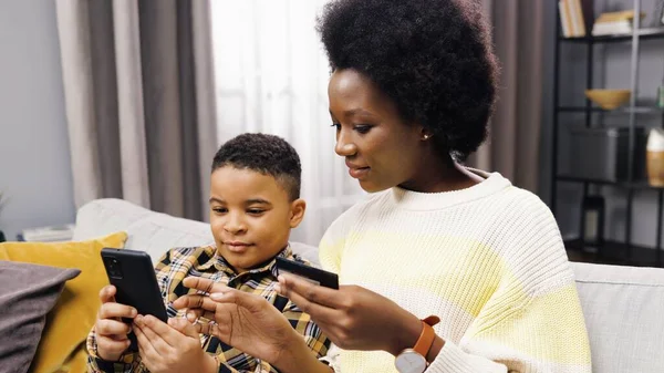 Genç ve güzel Afro-Amerikan annenin kanepeye oturup oğluyla banka kartı ve akıllı telefonu internetten alışveriş etmesi..