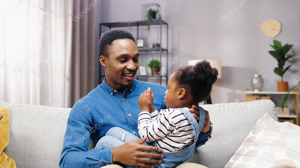 Happy young African American father sitting at home on couch with his cute little daughter and playing game with her while teaching counters.