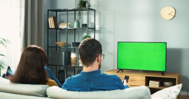 Genç bir çiftin arka planda televizyon izlerken kroma tuşlu kanepede dinlenirken program seçmeleri. Erkek ve kadın arkadaşlar uzaktan kumandayla televizyon izlerler.