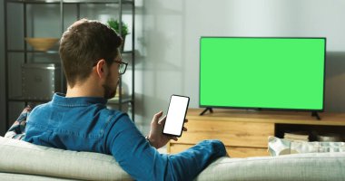 Gözlüklü, modern odadaki koltukta oturan, yeşil ekranlı televizyon izleyen ve internette krom tuşlu akıllı telefondan yazı yazan beyaz bir adam. Geçmişe bakış, boş zaman kavramı