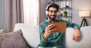 Evde oturma odasında dinlenirken, kulaklıkla film ya da tablet cihazıyla video izleyen Hintli genç adamın portresi. Yakışıklı sakallı adam internette aletleri karıştırıyor. Boş zaman konsepti