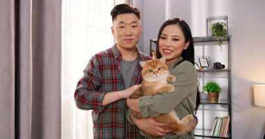 Neşeli, güzel, Asyalı eş ve kocanın evde durup sevimli kediyi ellerinde tutarak kameraya baktığı bir oda. Bakıcı sahipleri hayvan sever, aile üyesi konsepti