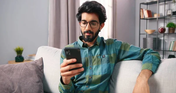 Close up of handsome young cheerful Hindu bearded guy sitting on couch at home in cozy living room and texting, browsing online on smartphone using social network app. Technology gadget