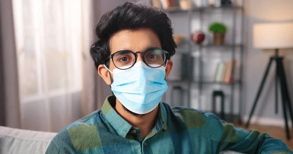 Neşeli Hindu sakallı yakışıklı adamın, sıcak oturma odasında oturmuş, karantinada, Coronavirus konseptinde gülümserken yüzündeki tıbbi maskeyi çıkarmasına yakından bakın. — Stok fotoğraf