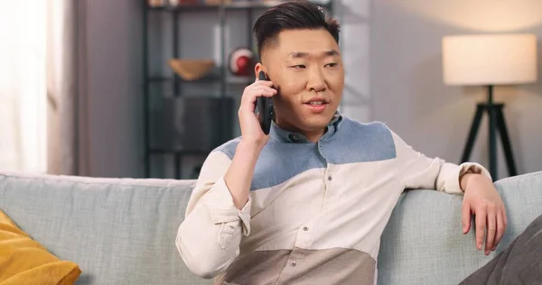 Портрет щасливого азіатського юнака, який сидить на дивані вдома і розмовляє по мобільному телефону. Хлопець, який кличе смартфон в доброму настрої, відпочиває в квартирі ізольовано один. Концепція розмови — стокове фото