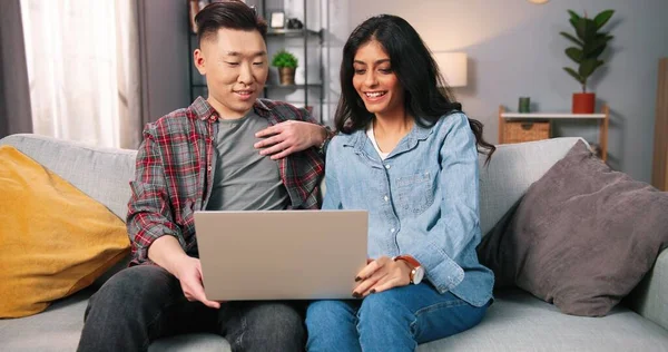 Joyful jong gemengd ras paar vriend en vriendin met behulp van laptop spreken, zoeken internet, tikken op de computer, praten op online videogesprek terwijl zitten op de bank in de moderne kamer, portret — Stockfoto