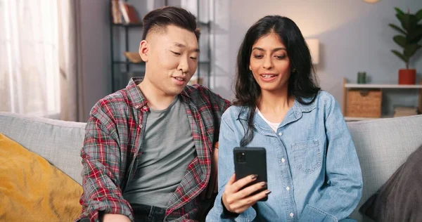 Portrait of multi-ethnic joyful happy couple wife and husband talking on video call online on smartphone while sitting on sofa in living room, videochatting, browsing on cellphone, leisure concept