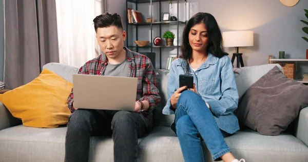 Multietniskt gift par sitter på soffan i mysigt vardagsrum hemma med hjälp av tech digitala prylar. Make bläddrar på bärbar dator medan hans fru söker på internet, sms på smartphone fritid koncept — Stockfoto