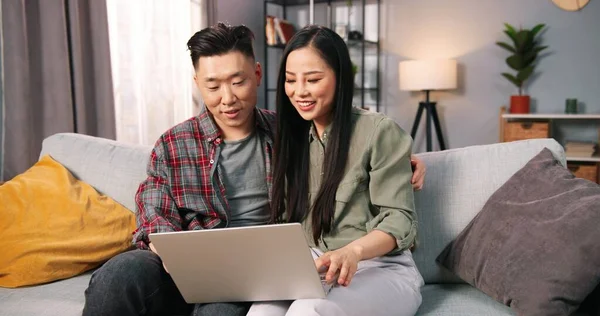 Trevlig asiatisk glad ung par fru och make ler och talar på gott humör videochattar online på laptop, bläddrar på datorn, talar spendera tid tillsammans på soffan hemma, porträtt — Stockfoto