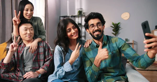 Retrato de la raza mixta feliz positivo joven Asiático e hindú parejas sentadas en el sofá en casa y posando sonriendo a la cámara del teléfono inteligente tomar fotos selfie juntos, fotos del teléfono celular, concepto de ocio —  Fotos de Stock