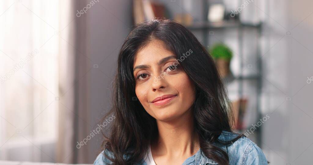 Close up of cheerful beautiful young Hindu female looking at camera and smiling in positive mood at home. Pretty dark-haired woman in good mood in living room, happy face, emotions concept