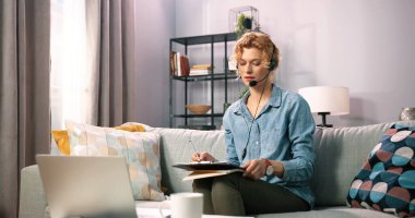 Kulaklık takan, koltukta oturan ve bilgisayarında çalışan odaklanmış bir kadın. Kulaklıklı kendine güvenen kız evde bilgisayar başında çalışıyor ya da önemli verileri kaydeden video semineri izliyor. Uzak çalışma