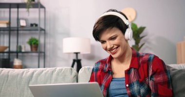 Gülümseyen kısa saçlı kadın, oturma odasında dizüstü bilgisayarıyla rahatlarken arkadaşıyla keyifli bir sohbetin tadını çıkarıyor. Kanepede oturan ve dizüstü bilgisayarda film izleyen kulaklık takan bir kadın.