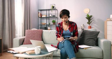 Kablosuz kulaklık takan gülümseyen bir kadın internetteki derste ve öğretmenlik yaparken bilgisayarını kullanıyor. Beyaz kadın öğretmen video konferans ile iletişim kuruyor evdeki kanepede oturuyor.