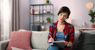 Kablosuz kulaklık takan gülümseyen bir kadın internetteki derste ve öğretmenlik yaparken bilgisayarını kullanıyor. Beyaz kadın öğretmen video konferans ile iletişim kuruyor evdeki kanepede oturuyor.