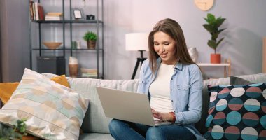 Kafkasyalı kahverengi saçlı kadın evdeki kanepede otururken dizüstü bilgisayarını kullanıyor. Milenyum kız öğrencisinin evden uzakta okuması için motive edilmiş. Bilgisayarda çalışan serbest çalışan kadın.