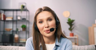 Kulaklıklı, mikrofonlu, kanepede oturan ve görüntülü konuşma yapan beyaz kadının portresi. Uzun saçlı kız iş arkadaşlarıyla online toplantı yapıyor.