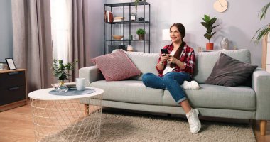 Sosyal ağlar. Beyaz tenli, esmer bir kadın evde akıllı telefonuyla mesajlaşırken pastel renkli koltukta rahatça oturuyor. Kız evde sohbet ediyor. Teknoloji ve insan konsepti. Stok fotoğrafı