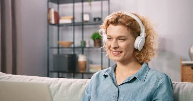 Gülümseyen kısa saçlı kadın, oturma odasında dizüstü bilgisayarıyla rahatlarken arkadaşıyla keyifli bir sohbetin tadını çıkarıyor. Kanepede oturan ve dizüstü bilgisayarda film izleyen kulaklık takan bir kadın.