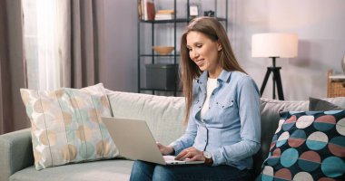 Kafkasyalı kahverengi saçlı kadın evdeki kanepede otururken dizüstü bilgisayarını kullanıyor. Milenyum kız öğrencisinin evden uzakta okuması için motive edilmiş. Bilgisayarda çalışan serbest çalışan kadın.