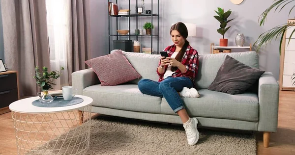 Sosyal ağlar. Beyaz tenli, esmer bir kadın evde akıllı telefonuyla mesajlaşırken pastel renkli koltukta rahatça oturuyor. Kız evde sohbet ediyor. Teknoloji ve insan konsepti. Stok fotoğrafı