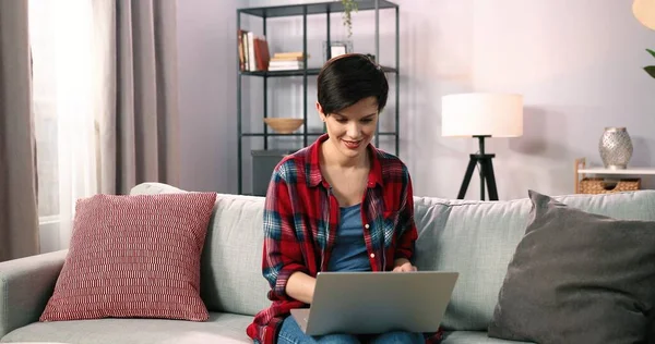 Evdeki rahat koltukta dizlerinin üstüne çöküp internette gezinen gülümseyen bir kadın. Mutlu kısa saçlı kız haftasonunu evde geçirirken koltukta dinleniyor ve bilgisayarda film seyrediyor.