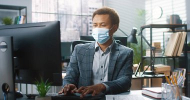 Tıbbi maske takan genç Afrikalı Amerikalı erkek işçinin portresi iş yerinde öksürürken ve baş ağrısı çekerken bilgisayarda yazıyor. Coronavirus salgını, covid-19 semptomları. Çalışma konsepti